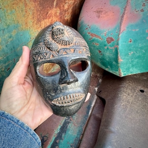 May include: A dark, carved mask with an oval shape and eye and mouth openings. The mask features a raised snake design on top and intricate patterns around the forehead. The mask is held in a hand against a backdrop of weathered metal.