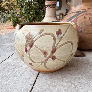 May include: A beige ceramic bowl with a rounded shape and a wide opening. The bowl is decorated with brown and red floral patterns. The bowl is sitting on a wooden surface, with other ceramic pieces in the background.