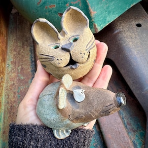 May include: Two handmade ceramic figurines: a smiling cat head and a mouse. The cat is tan with black stripes and green eyes. The mouse is brown and gray with a black nose and a white ear. The figurines are held in a hand.