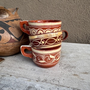 May include: A stack of three terracotta-colored ceramic mugs with cream-colored hand-painted geometric and floral patterns. Each mug features a sturdy handle and a wide rim. Beside the mugs sits a large, matching terracotta-colored ceramic pitcher.