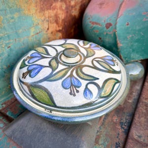 May include: A ceramic lidded dish with a floral design in blue, green, and tan. The lid has a small knob and a handle on the side. The dish is round and has a blue rim.