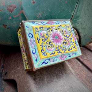 May include: Rectangular antique box with a yellow background and floral design. The box features a pink flower in the center, surrounded by blue and green accents. The edges are trimmed with a brown metal.