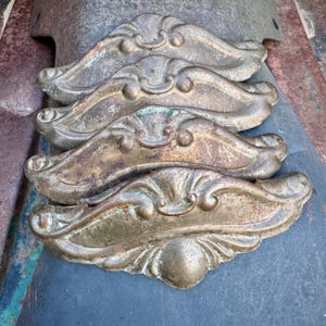 May include: Four antique brass decorative architectural elements. Each element features an ornate, symmetrical design with scrollwork and a central, rounded detail. The brass has a weathered, aged patina with hints of green and brown.