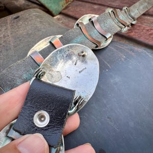 May include: Close-up of a vintage belt with a silver buckle and black leather strap. The buckle has an oval shape with intricate metalwork and the inscription "Sterling" and "J.N.". The belt's leather is a weathered gray color.