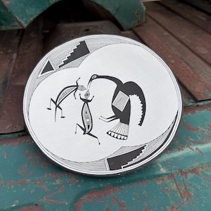 May include: A round, white ceramic plate with a black tribal design. The design features stylized figures and a bird. The plate has a black rim and is set against a weathered, green surface.