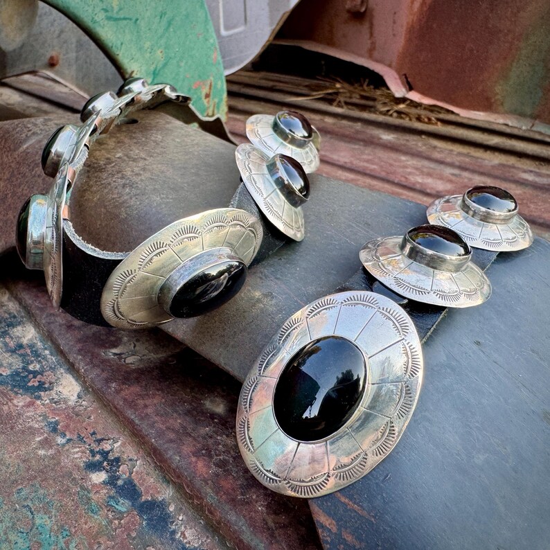 May include: A silver belt buckle set with black oval stones. The set includes a belt buckle and several belt loops, all with a decorative, engraved design. The black stones are set in a silver metal. The belt is black.