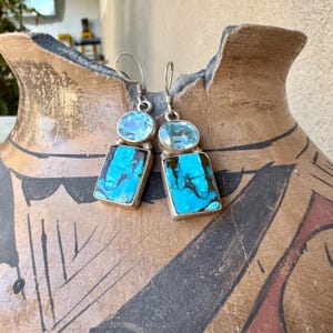 May include: A pair of silver earrings with a turquoise and blue gemstone design. Each earring features a rectangular turquoise stone below a smaller, oval-shaped blue stone. The earrings are displayed against a textured, brown background.