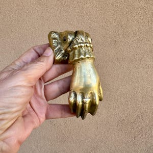 以下が含まれることがあります： 手の形をした真鍮製のドアノッカー。手には関節のディテールとフリルの袖口があります。ノッカーは温かみのある金色で、無垢の金属でできているようです。