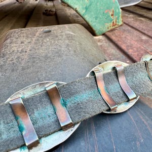 May include: Close-up of a vintage belt with a dark gray leather strap and large, oval silver-toned buckles. The belt features copper-colored metal clips and shows signs of wear, with a weathered appearance.