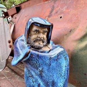 May include: A ceramic statue of a bearded man wearing a blue hooded robe. The statue has a detailed face with a stern expression. The robe has a textured, speckled pattern in shades of blue.