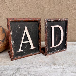 May include: Two vintage-style metal signs, each with a letter. One sign displays the letter "A", the other displays the letter "D". The letters are off-white against a black background, framed in a distressed, reddish-brown metal.