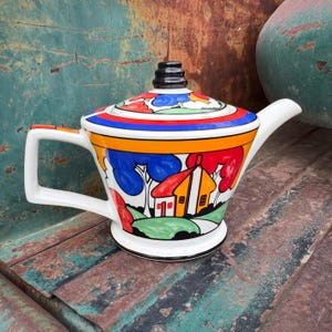 May include: A white teapot with a black knob on the lid. The teapot is decorated with a colorful art deco design of a house, trees, and hills. The teapot is sitting on a rusty metal surface.