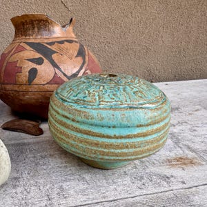 May include: A low, rounded ceramic vessel with a turquoise and brown glazed finish, featuring horizontal ridges along the sides and a textured, patterned top. Beside it sits a larger, bulbous terracotta pot with black and red geometric designs.