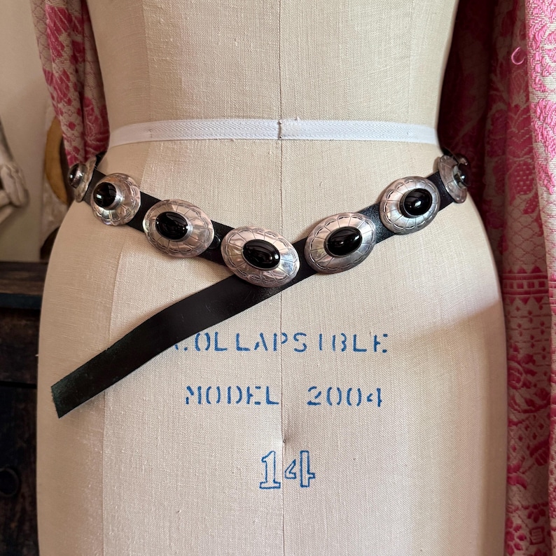 May include: A black leather belt with silver-toned oval conchos, each featuring a black stone center. The belt is displayed on a mannequin. The conchos have a detailed, embossed design. The mannequin is marked with the text "COLLAPSIBLE" and "MODEL 2004".
