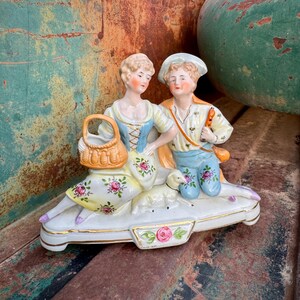 May include: A porcelain figurine of a couple sitting on a white base. The woman is holding a basket and the man is holding a pipe. The figurine is decorated with floral patterns and has a gold trim.