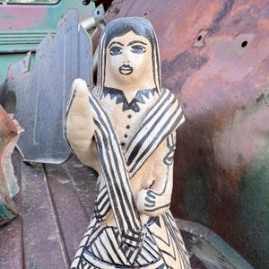 May include: A hand-painted ceramic figurine of a woman with black hair and a black and white striped shawl. The figurine has a cream-colored base with black details on the dress and arms. The figure is standing in front of a rusty metal background.