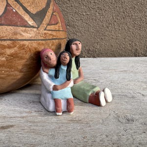 May include: A handcrafted ceramic sculpture of three figures. One figure embraces a child in a blue dress, while another sits nearby in a green dress. The figures have dark hair and are set against a rustic background.