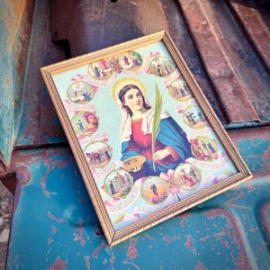 May include: Framed religious artwork featuring a central portrait of a woman with a halo, holding a palm frond and a plate. The artwork is surrounded by smaller circular illustrations and framed in gold.