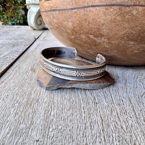 May include: A silver cuff bracelet with an open design, resting on a small, dark stone. The bracelet features an engraved pattern of oval and star-like shapes. A large, brown, ceramic bowl is in the background, along with a weathered wooden surface.