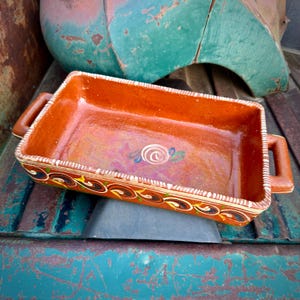 May include: A rectangular, terracotta-colored baking dish with two handles. The rim is decorated with a repeating pattern of brown, yellow, and white swirls. The interior features a painted spiral design with blue and green accents.