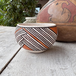 May include: A small, round ceramic bowl with a white base and a pattern of black, white, and orange diagonal stripes. The bowl is set on a weathered wooden surface, with a larger, decorated ceramic vessel in the background.