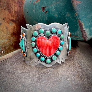 May include: A wide silver cuff bracelet featuring a large central heart-shaped red stone surrounded by a border of small round turquoise cabochons. The silver metal surface is decorated with etched line patterns and additional turquoise accents.