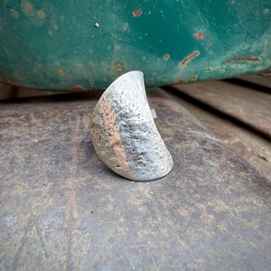 May include: A silver-toned ring with a textured, crescent-shaped design. The ring has a rough, pitted surface with a band of copper-colored metal. The ring is set against a weathered, gray surface.