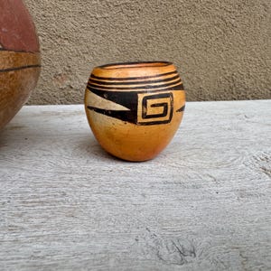 May include: A small, hand-painted ceramic pot with a rounded shape. The pot features a warm orange base with black geometric designs, including a square spiral and horizontal stripes near the rim. The pot is displayed on a weathered wooden surface.