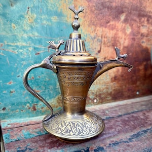 Puede incluir: Una tetera de latón vintage con intrincados patrones grabados y adornos en forma de pájaro. La tetera presenta un asa curva y un pico largo, con una figura de pájaro en la tapa y dos más en el pico. El latón tiene una pátina cálida y envejecida.