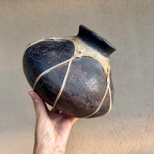 May include: A dark brown, rounded ceramic vessel with a wide, short neck. The pot features a textured, weathered surface and is secured by a network of light-colored, thin cordage that wraps around the body and connects near the rim.