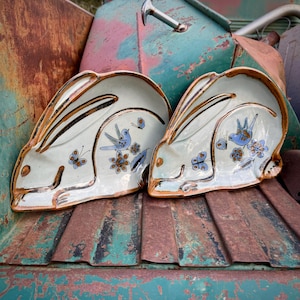 May include: Two ceramic rabbit-shaped plates. The plates are off-white with brown and black accents. Each plate features a blue bird, butterflies, and floral designs. The plates are displayed on a weathered, rusty metal surface.