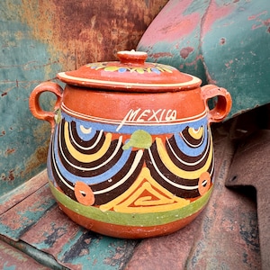 May include: A terracotta-colored ceramic pot with a lid, featuring hand-painted designs in blue, yellow, black, and green. The word "MEXICO" is written on the side. The pot has two handles and a small knob on the lid.