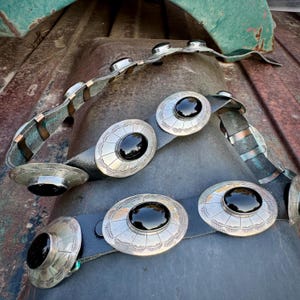 May include: A black leather belt with large, oval silver buckles, each featuring a black stone center. The silver buckles have an etched design. The belt is displayed on a weathered, rusty metal surface.