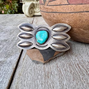May include: A silver-toned belt buckle with a turquoise stone center. The buckle has an ornate design with oval shapes and a scalloped edge. The turquoise stone is a vibrant blue-green color. The buckle is likely vintage or antique.