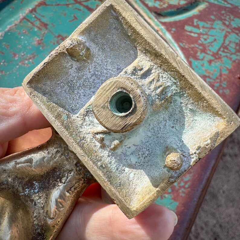 以下が含まれることがあります： ヴィンテージの真鍮製オブジェクトの下面のクローズアップ。おそらく家具の部品です。正方形のベースには、中央にねじ穴と2つの小さな突起があります。真鍮は、緑青と摩耗による経年変化を示しています。