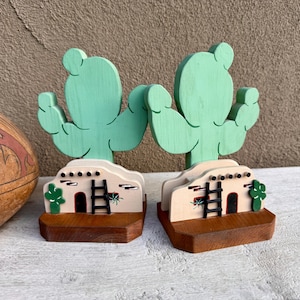 May include: Two wooden cactus bookends. The cacti are painted light green and sit atop a tan-colored base with a Southwestern design. The base is mounted on a dark brown wooden block.