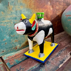 May include: A hand-painted wooden cow figurine with black and white spots. The cow has yellow horns, red and green details, and a green and white hat. The cow is standing on a yellow and blue base.