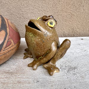 May include: A ceramic frog sculpture with a wide open mouth and yellow eyes. The frog is brown with green accents and sits on a wooden surface. The sculpture is next to a brown and red patterned vase.