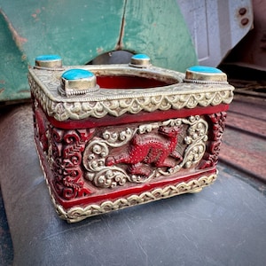 May include: A square, red and silver decorative box with turquoise accents. The box features intricate carvings of a dragon and floral patterns. The top has four turquoise stones set in silver. The box is likely made of wood and metal.