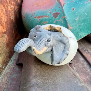 May include: A gray elephant emerging from a cracked, off-white egg. The elephant has detailed features, including a trunk, ears, and eyes. The egg is set against a backdrop of weathered metal in shades of brown, red, and green.
