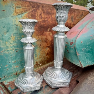 May include: Two silver-colored, ornate candle holders. The candle holders have a fluted column design with decorative leaf accents. The bases are hexagonal with detailed patterns. The tops are shaped like small urns with leaf patterns.