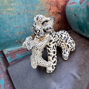 May include: A ceramic sculpture featuring a large leopard with black spots and stripes on a cream-colored base, depicted in a playful pose with a smaller leopard cub. The figures are hand-painted with intricate black markings and detailed facial features.