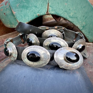 May include: A black leather belt with large, ornate silver buckles. Each buckle features an oval, polished black stone set within a detailed, engraved silver frame. The belt is displayed against a weathered, vintage background.