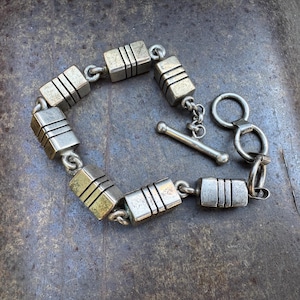 May include: Silver-toned bracelet featuring rectangular links with three horizontal lines. The bracelet has a toggle clasp and is displayed on a textured, dark surface. A piece of jewelry.