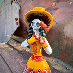 May include: A Day of the Dead figurine of a female skeleton wearing a yellow dress and hat. The figurine holds a bouquet of red flowers. The dress has a red and white geometric pattern on the skirt and a decorative design on the bodice.