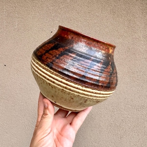 May include: A handmade ceramic bowl with a unique design. The bowl features a combination of brown, black, and beige colors. The exterior has horizontal stripes and a textured surface. The bowl is held in a hand.