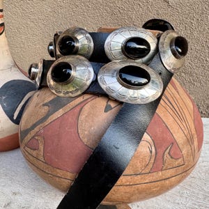 May include: A black leather belt with silver-toned oval conchos, each featuring a black stone center. The belt is displayed over a decorative, hand-painted pottery vessel with red and brown geometric designs. The conchos are approximately 2 inches in diameter.