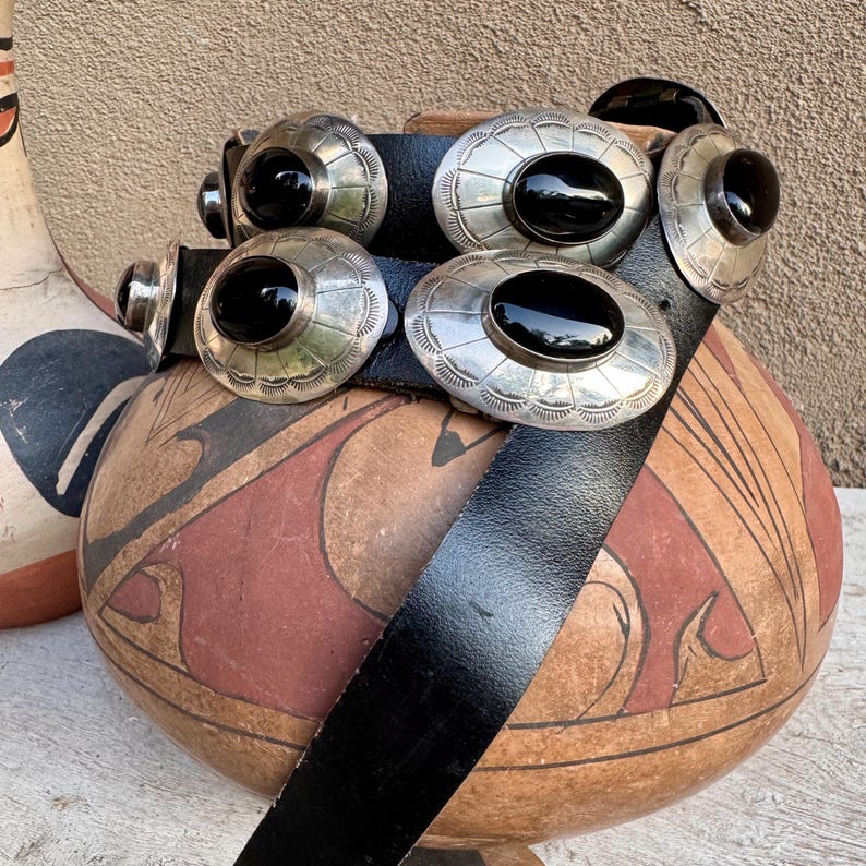 May include: A black leather belt with silver-toned oval conchos, each featuring a black stone center. The belt is displayed over a decorative, hand-painted pottery vessel with red and brown geometric designs. The conchos are approximately 2 inches in diameter.