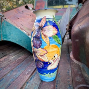 May include: A tall, hand-painted ceramic vase with a floral and swan design. The vase features a blue background with yellow and purple flowers, a yellow sun, and two white swans. The top and bottom rims are cream and blue.