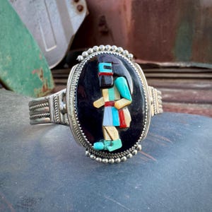 May include: A silver bracelet with a black oval inlay featuring a colorful mosaic of a Native American figure. The bracelet has a beaded edge and a braided band.